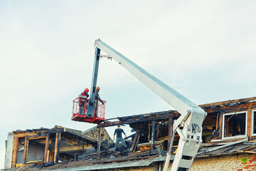 Fototapeta premium Unrecognizable construction team operating crane to dismantle charred roof in the aftermath of fire. Post-fire dismantling of a burnt roof with a crane cradle and team of workers