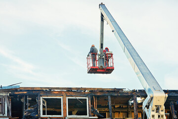 Workers on crane cradle dismantle burnt roof after fire, team effort in the cleanup and reconstruction process.