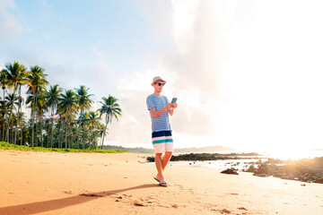 Brazilian man standing on the sand of a paradise beach. He is using his smartphone, either to send a message or use an app.  In the background, coconut trees and the light of the sunrise.