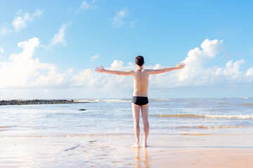 Brazilian man wearing swimwear, with his arms raised to express gratitude and happiness for enjoying summer vacation on the beach. He is looking at sea.