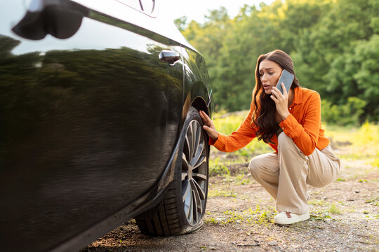 Worried young European woman talking on cellphone and checking her car flat tire on the side of the highway, calling for help