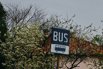 Fototapeta premium Bus stop sign in village. House with a tiled roof in Balkans. Beautiful apple tree or cherry plum with white flowers. Bijeljina is a small town on the border with Serbia and Bosnia and Herzegovina.