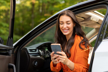 Cheerful young European lady using modern cellphone and sitting in car with opened door on sunny...