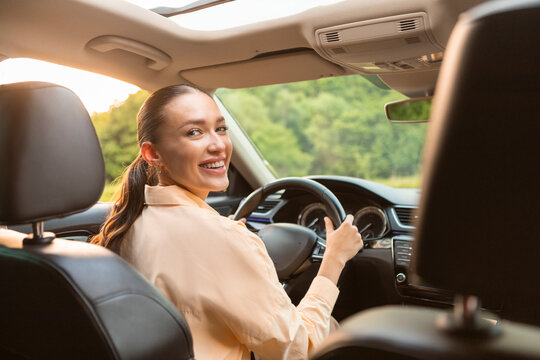 Happy woman driving luxury car, sitting on drivers seat and smiling looking back, testing her newest automobile, holding steering wheel