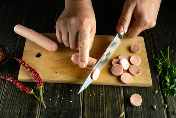 Slicing sausages on a kitchen board with a knife in the hand of a chef to prepare a delicious dietary dish for breakfast.