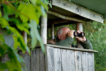 Older, experienced hunter sitting on his hunting pulpit and looking through his binoculars.