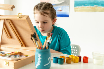Portrait of a little girl artist, she looks with a concentrated gaze at an easel, there are many...