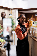 Young black woman records a video in a zero-waste shop promoting fresh, organic products. She uses...
