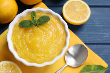 Delicious lemon curd in bowl, fresh citrus fruits and spoon on blue wooden table, flat lay