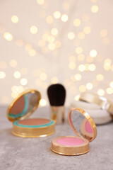 Face blusher and other cosmetic products on grey textured table against blurred lights, closeup....