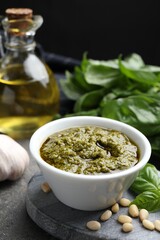 Tasty pesto sauce in bowl, basil, pine nuts, garlic and oil on grey table
