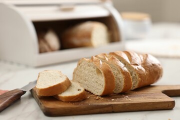 Wooden bread basket with freshly baked loaves and knife on white marble table in kitchen, closeup