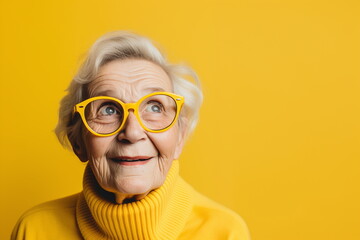 A cheerful elderly woman sporting vibrant yellow glasses and a turtleneck sweater looks upwards with a joyful expression, standing before a matching yellow backdrop - Generative AI