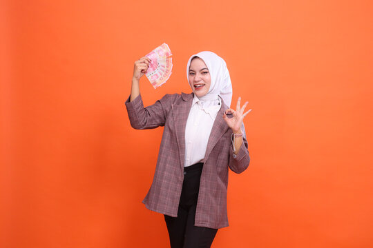 Excited beautiful Asian girl wearing hijab cheerful holding rupiah banknotes and signing okey (steady good) isolated on orange background. finance, lifestyle, advertising concept