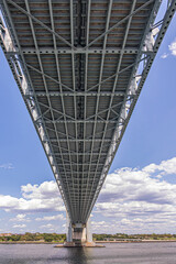 New York, NY, USA - August 1, 2023: Right under Verrazzano-Narrows Bridge E side half at Fort Hamilton. Green coastline under blue cloudscape