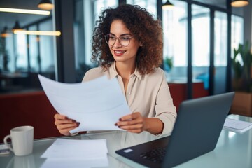 Young happy and successful businesswoman in glasses working with documents inside office, Hispanic woman with laptop looking at bills and contracts, financier with curly hair using laptop Young happy 