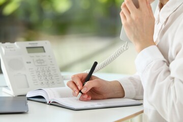 Assistant with telephone handset writing at white table against blurred green background, closeup