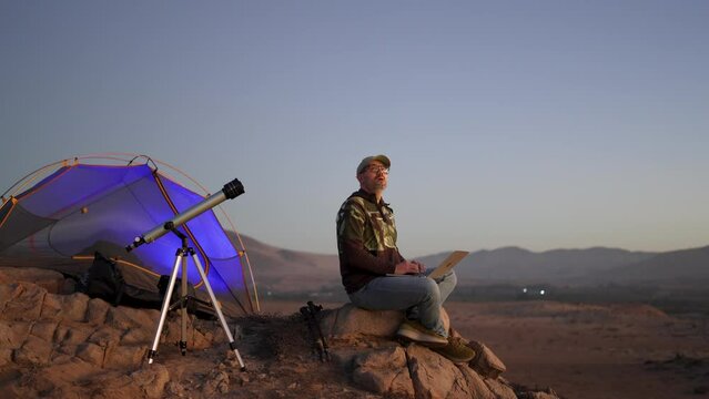 digital nomad man sitting alone with a laptop in the desert at blue our next to a telescope watching the sky