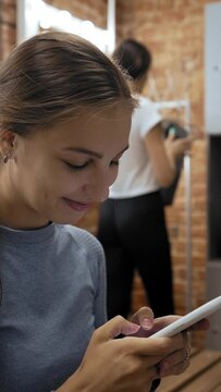 Focused woman texts with boyfriend via smartphone while girl friend opens locker and puts clothes. Ladies in dressing room before training closeup Vertical Shot.