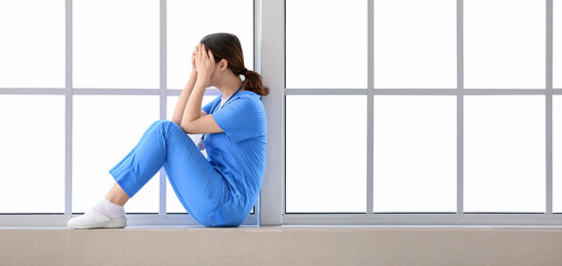 Fototapeta premium Depressed Asian doctor sitting on window sill in clinic