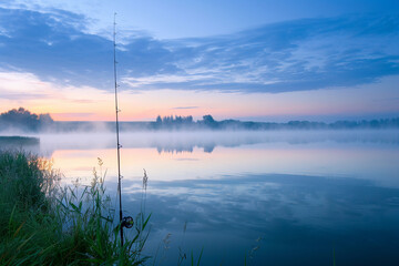 A lakeside fishing scene at dawn, with a lone fishing rod propped beside a calm, misty lake.