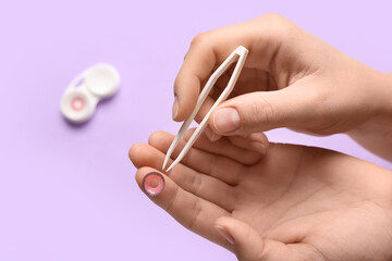 Female hands with tweezers and container with contact lenses on lilac background