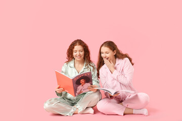 Happy redhead sisters in pajamas with magazines on pink background