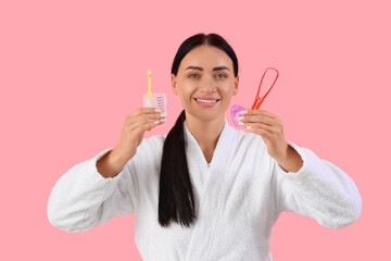 Beautiful young happy woman in bathrobe with supplies for oral hygiene on pink background