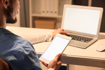 Male nurse with tablet computer and laptop working evening shift at table in clinic, closeup