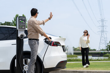 Young couple recharge EV car battery at charging station connected to power grid tower electrical...