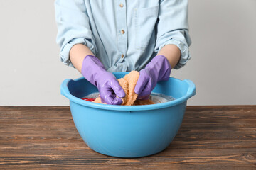 Woman in rubber gloves washing clothes in plastic basin on wooden table against light background