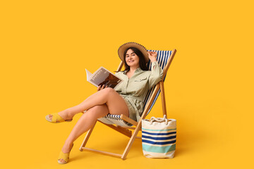 Beautiful young woman with magazine and stylish bag sitting in deck chair on yellow background