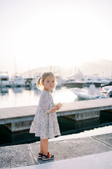 Little girl stands on the pier, moving her hands and looking to the side