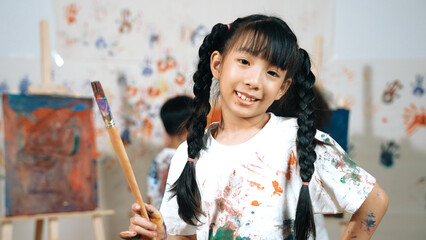 Creative cute girl pose at camera while diverse children painting behind. Young beautiful child...