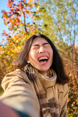Young Asian man with long hair laughing and taking a funny selfie in a park in autumn.