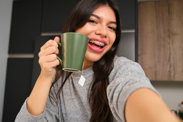 Happy female teenager with braces and a cup of tea taking a selfie in the kitchen.