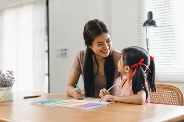 Young mother assists her daughter with coloring activities at a home desk during daytime.