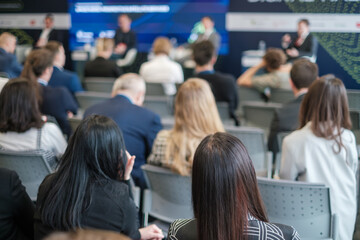 Audience members sitting and listening attentively to speakers at a business conference. People are engaged in the presentation, seated in rows facing the stage.