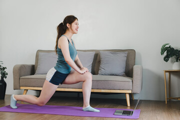 Young Asian woman at home doing yoga in front of open laptop, repeating instructions by professional online fitness trainer.