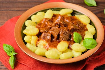Triestine beef goulash in tomato and sweet paprika sauce, served with potato gnocchi, in a ceramic plate on a brown wooden background. Beef recipes.