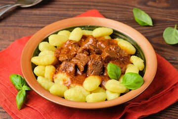 Triestine beef goulash in tomato and sweet paprika sauce, served with potato gnocchi, in a ceramic plate on a brown wooden background. Beef recipes.