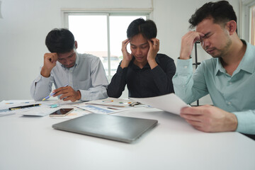 A team of tired, stressed young businessmen hold their heads in their hands. From working with problems without success Financial documents in the Ahimsarimsap project Office syndrome concept.