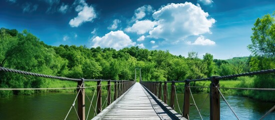 A wooden suspension bridge spans a river in a vibrant summer setting surrounded by lush green trees...