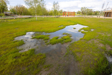 A serene wetland scene within an urban park, featuring a reflective pond, lush green grass, and a wooden bridge leading to a striking red structure.