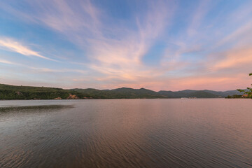 A serene sunset over a tranquil lake with rippled water, mountains in the distance, and a clear sky with soft clouds...