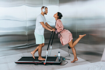woman in a robe kisses a man on a treadmill. The man is wearing headphones and exercise clothes, highlighting a mix of romance and fitness