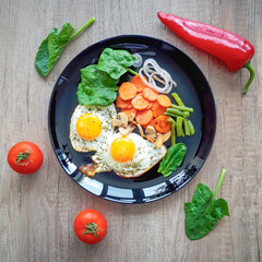 Fried eggs, carrots, spinach, green beans on a black plate. Red peppers and tomatoes lie nearby. Healthy breakfast. Bright vegetables on a textured background. View from above