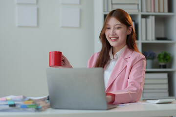 Asian businesswoman sitting, relaxing, holding a hot coffee mug, working with laptop on table, online finance, documents, taxes, reports, marketing, statistics, business analysis research concept.