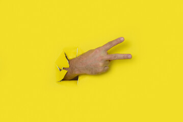 Hand making a sign of victory, peace, coming out of the hole in a torn yellow paper background.