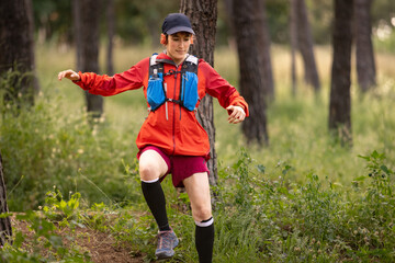 A woman in a red jacket and red shorts is running through a forest. She is wearing a black hat and...